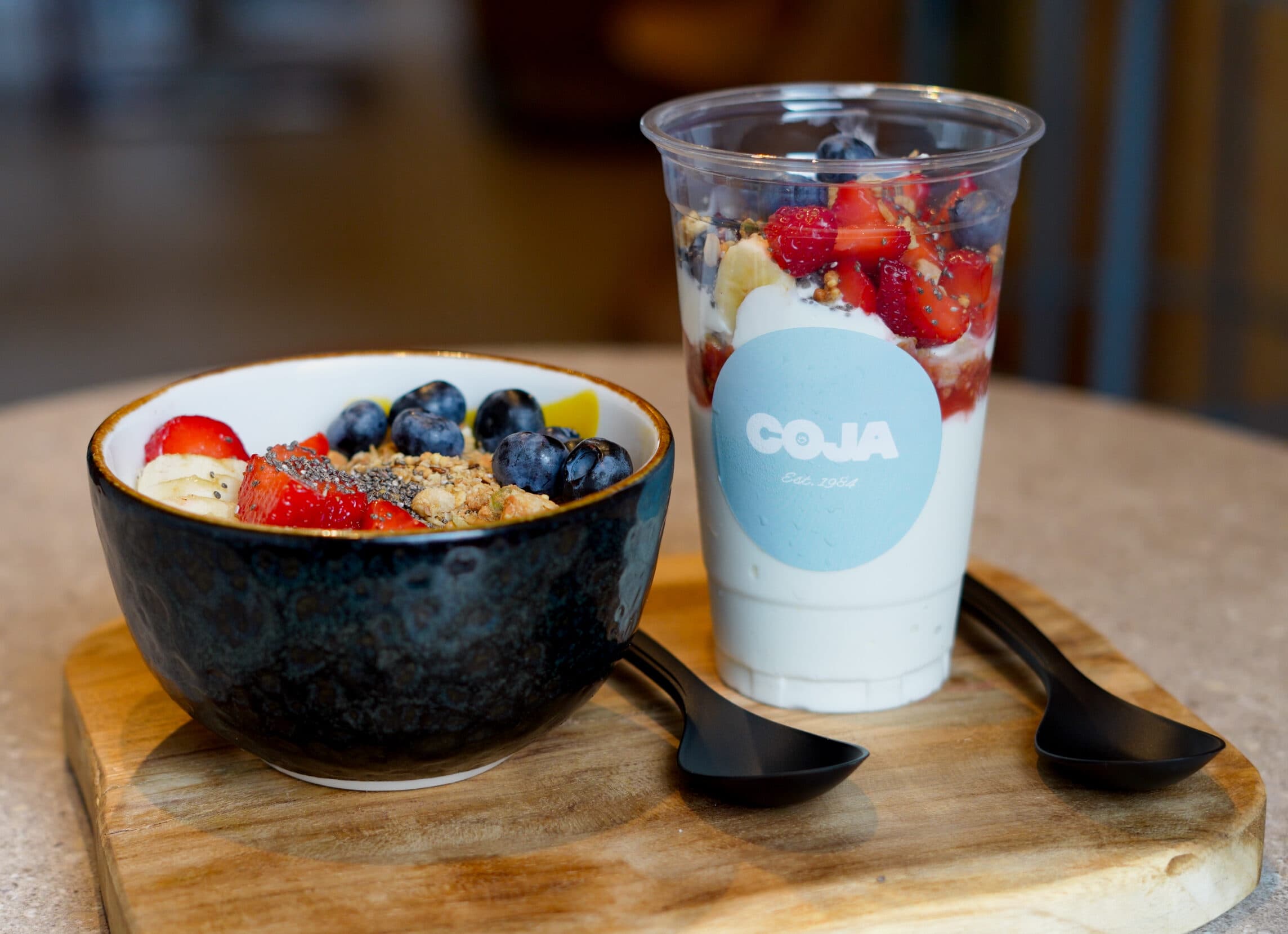 Yoghurtbowl met granola, aardbeien, blauwe bessen, banaan en chiazaad, naast een Coja-beker met yoghurt en fruit.