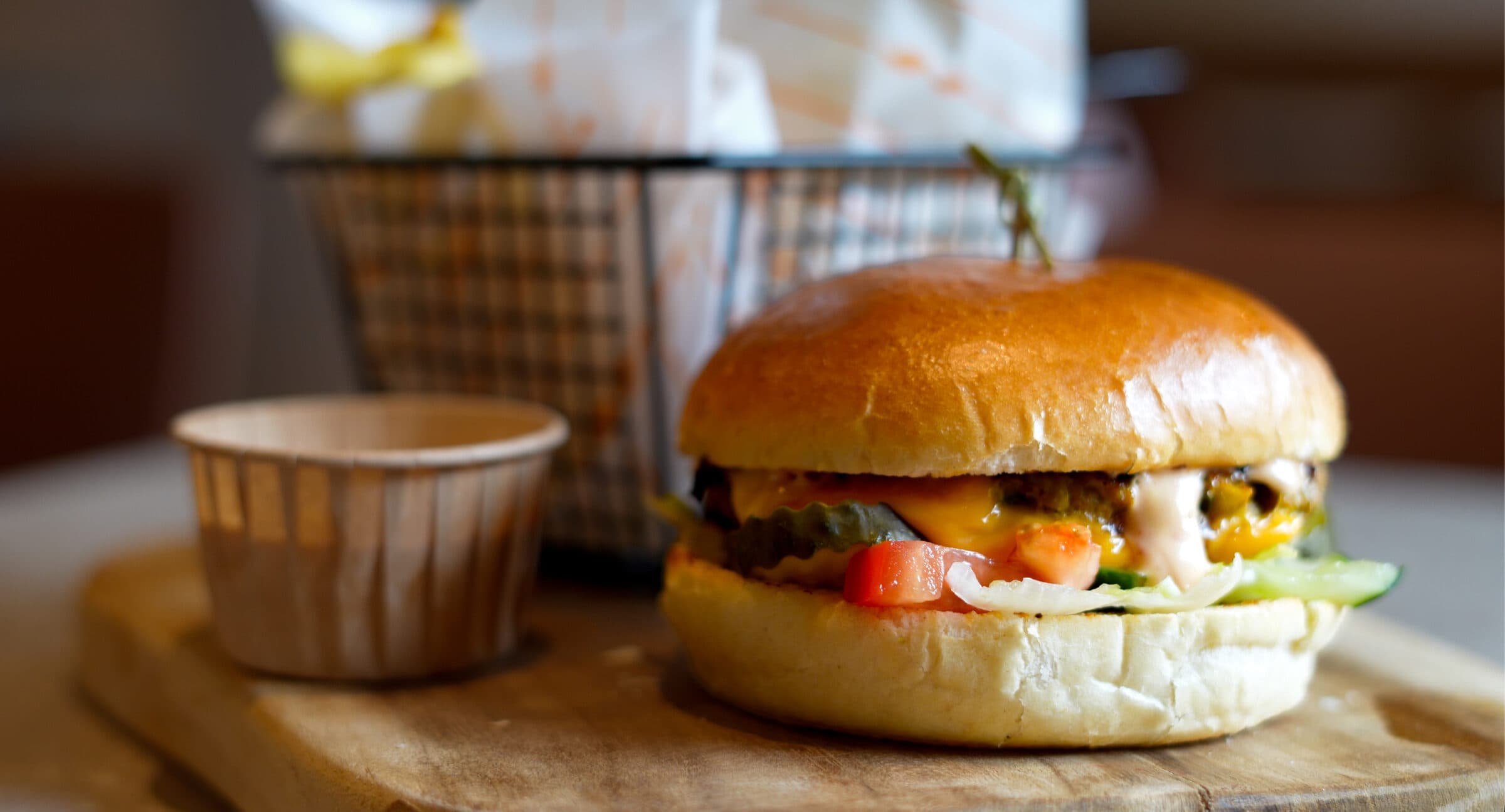 Hamburger op een briochebroodje met cheddar, tomaat, augurk en sla op een houten plank, met een mandje patat erachter.