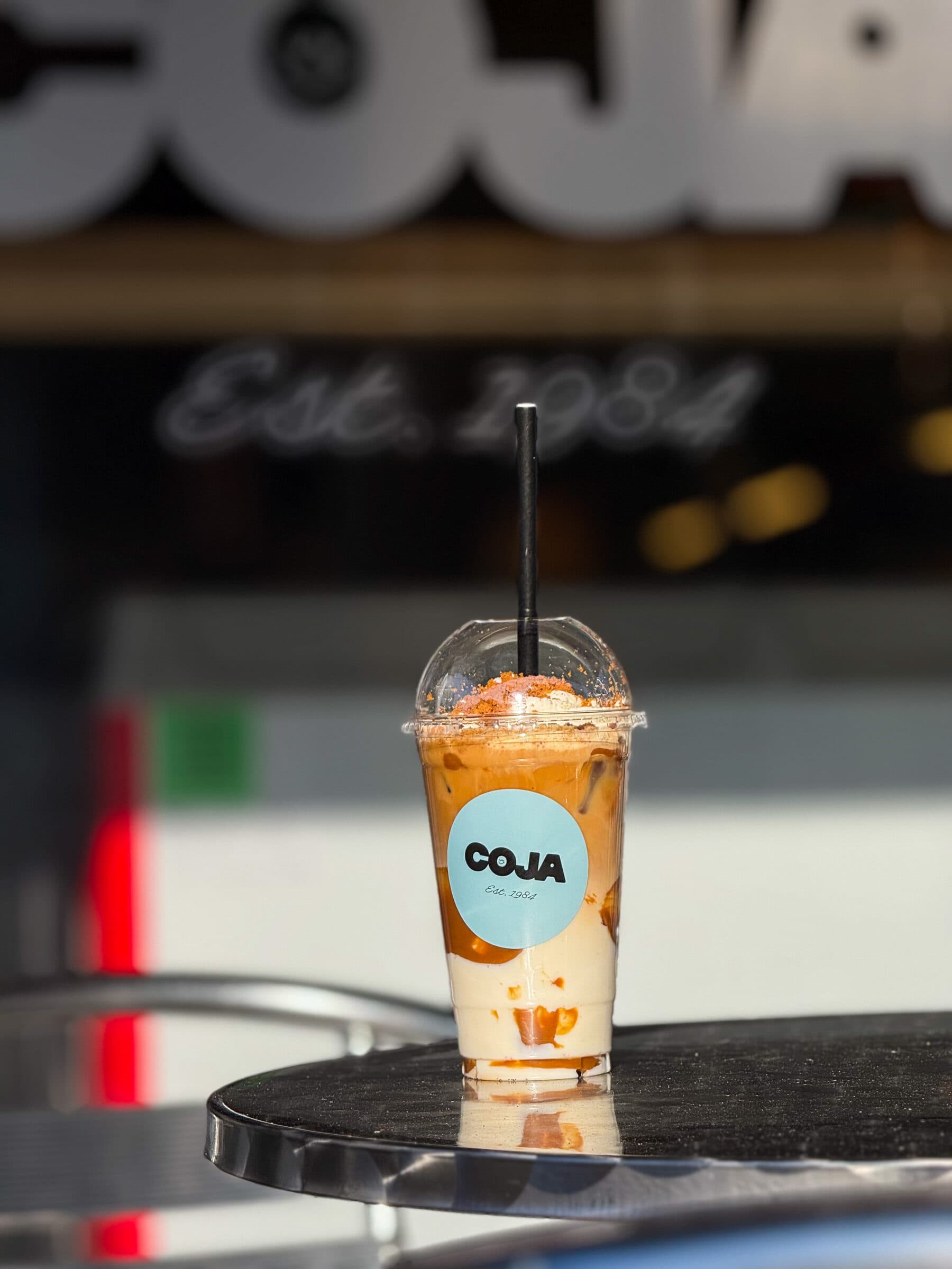 IJslatte macchiato in een Coja-beker met rietje op een terrastafeltje, winkelpui op de achtergrond.