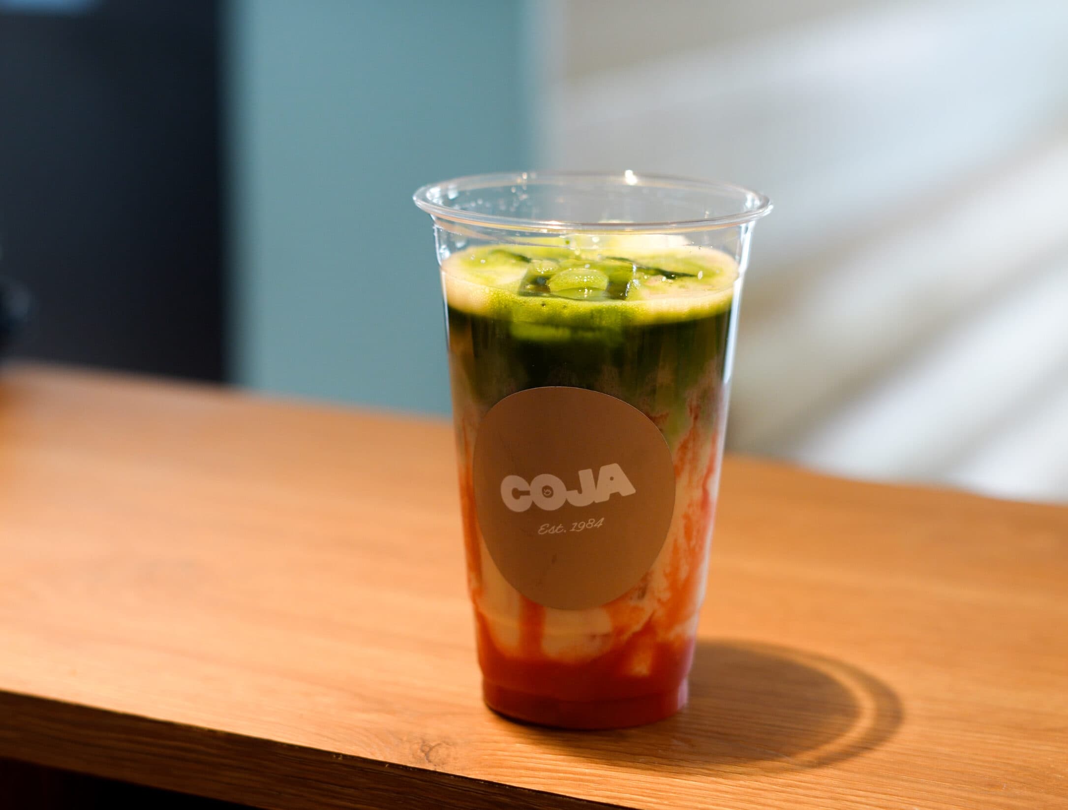 IJsmatcha in een Coja-beker, gelaagd met aardbeiensiroop, melk en groene matcha, op een houten tafel.
