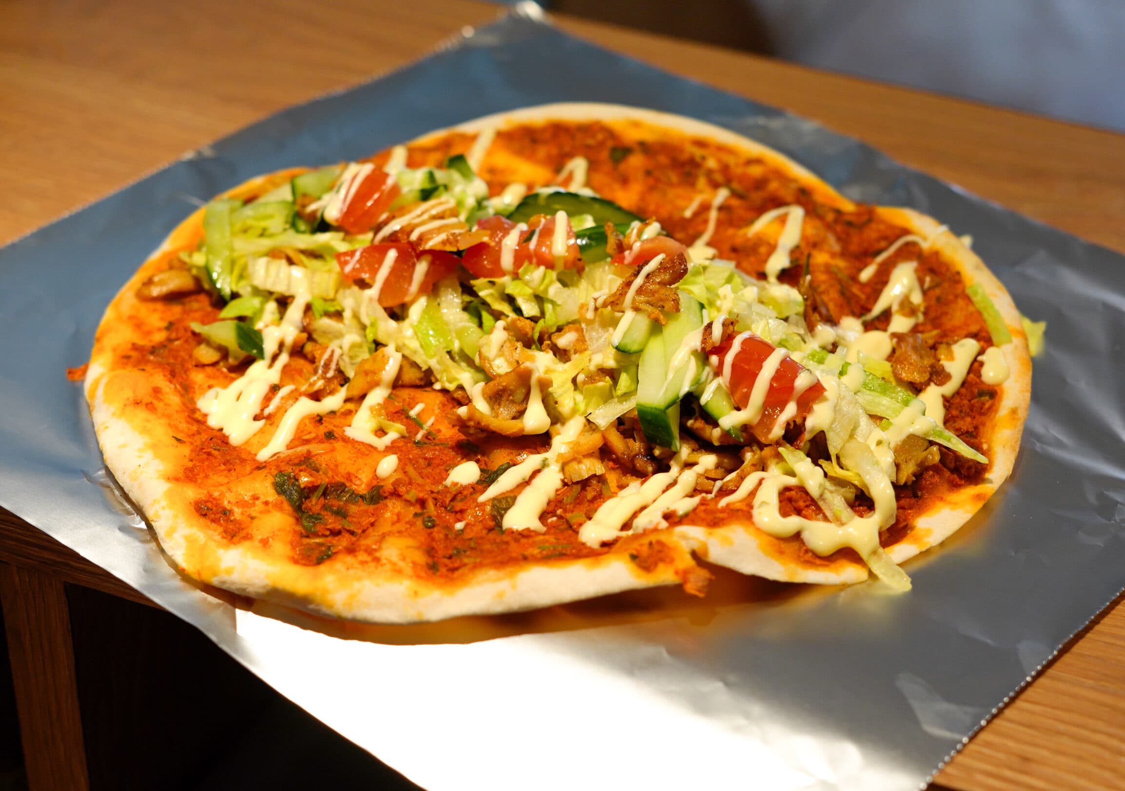 Turkse pizza met shoarma, sla, tomaat, komkommer en knoflooksaus, uitgevouwen op folie.