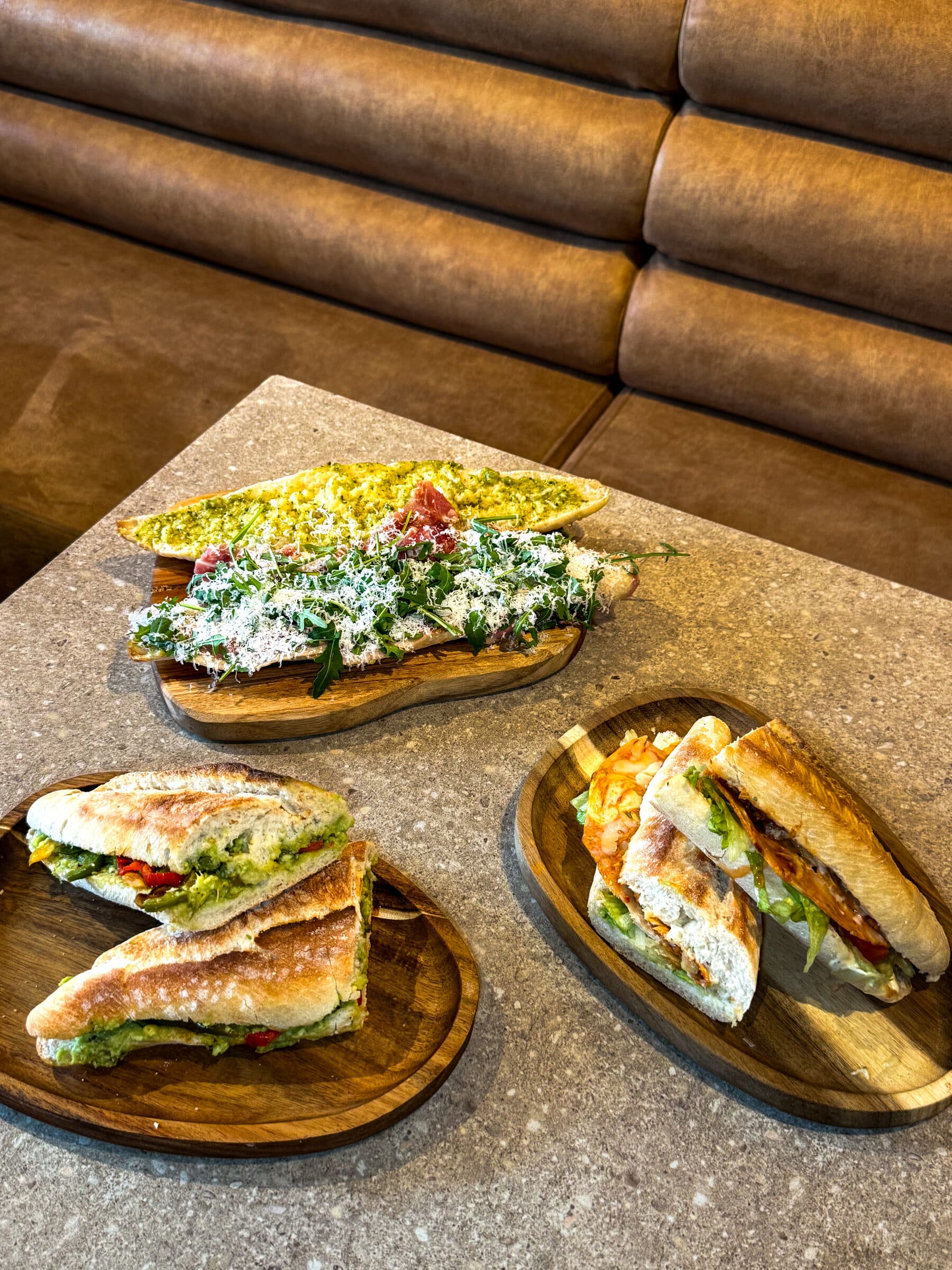 Drie broodjes op houten plankjes: een open broodje met rucola en Parmezaan en twee geperste broodjes, op een bruine bank.