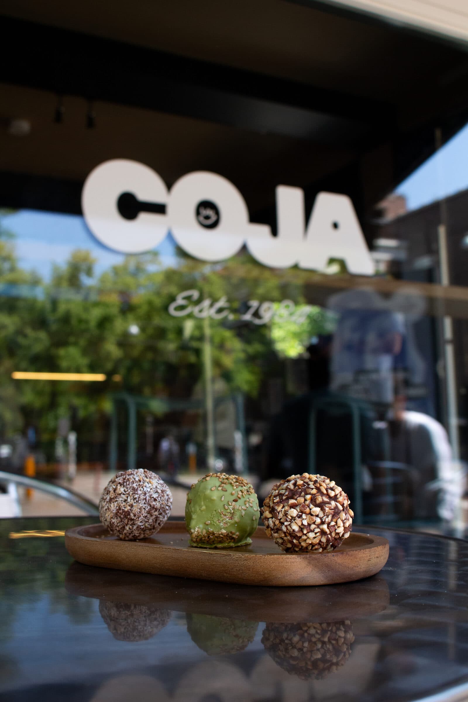 Drie energieballen met kokos, matcha en hazelnoot op een houten schaaltje voor de etalage met het COJA-logo.