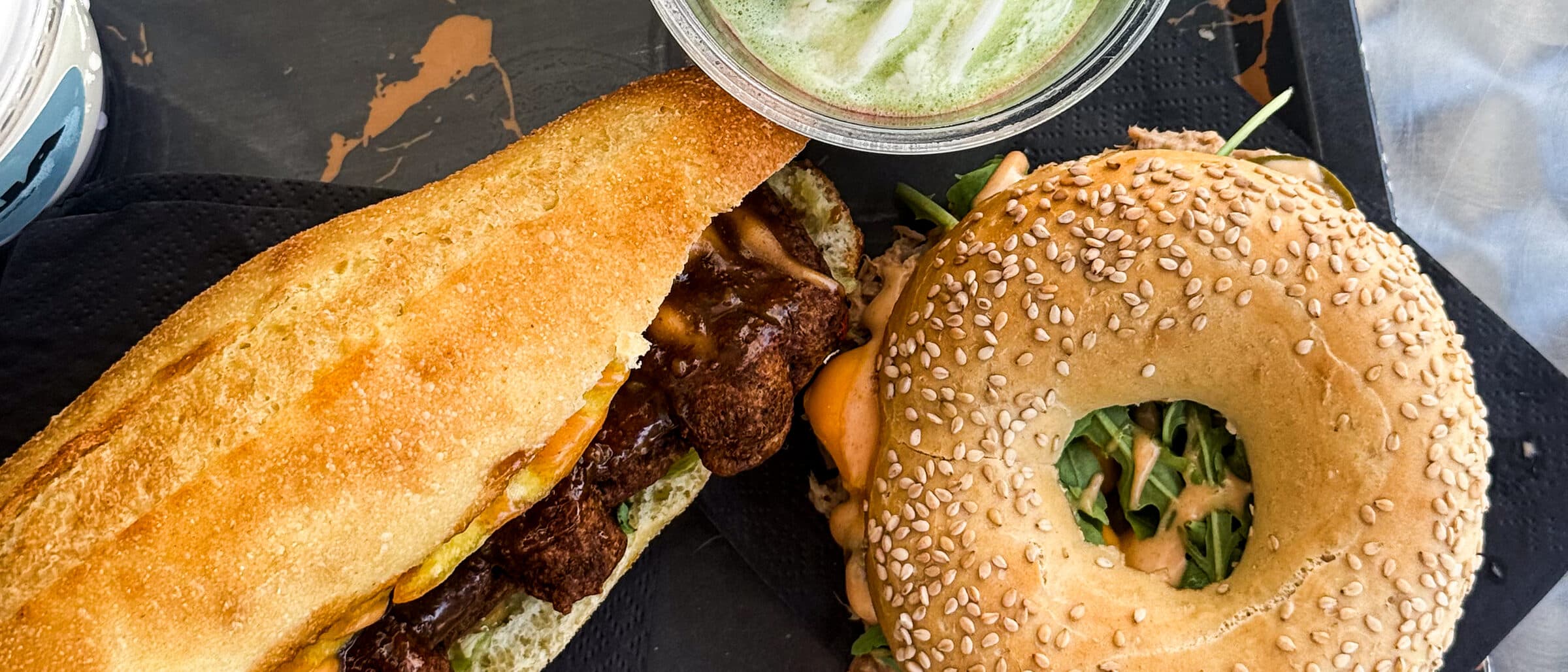 Dienblad van bovenaf met een broodje, een sesambagel met tonijn en rucola en een matcha latte op zwarte servetten.