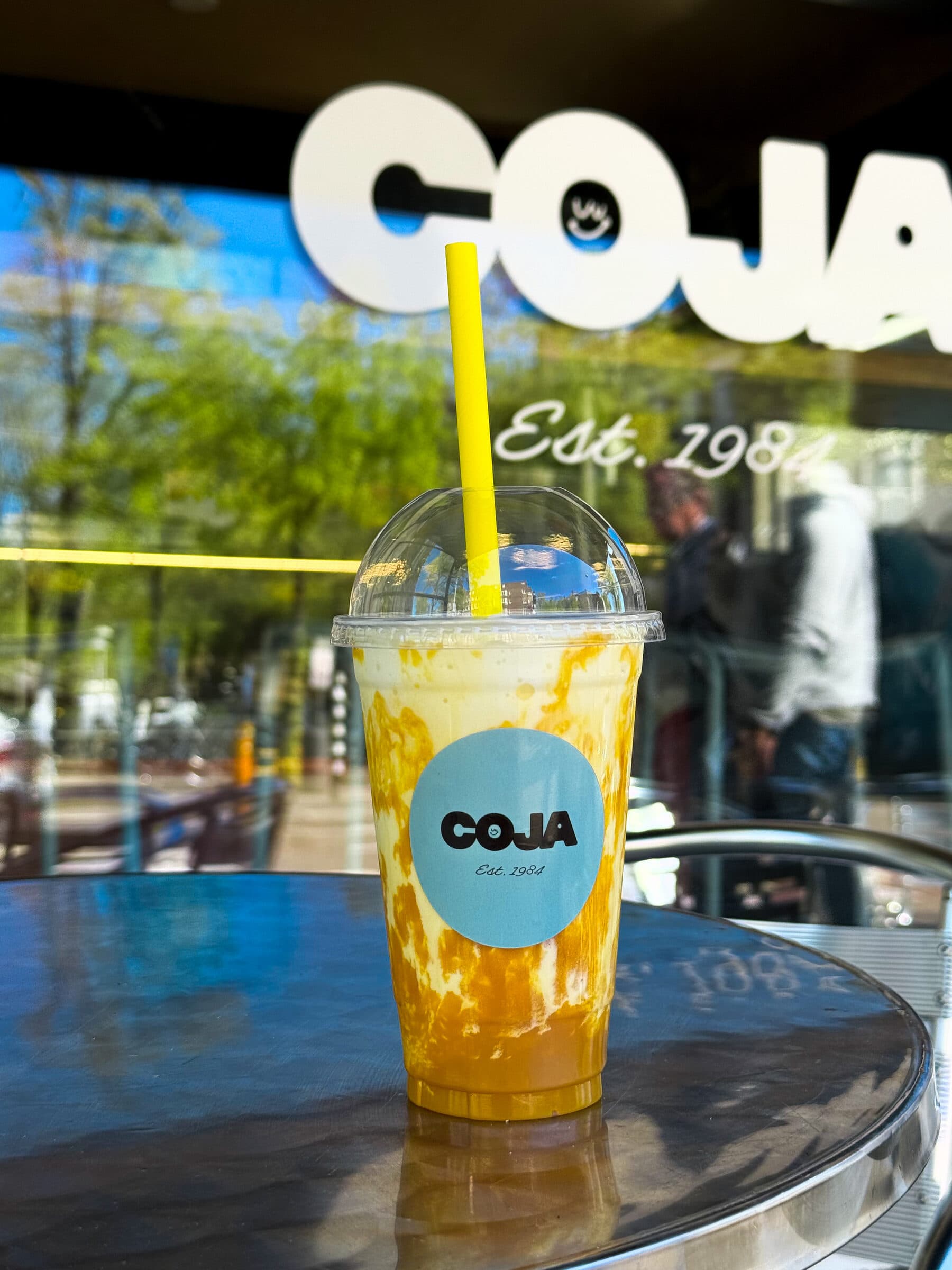 Mango smoothie in een beker met geel rietje op een tafel bij het raam, met het Coja-logo en 'Est. 1984' op het glas.